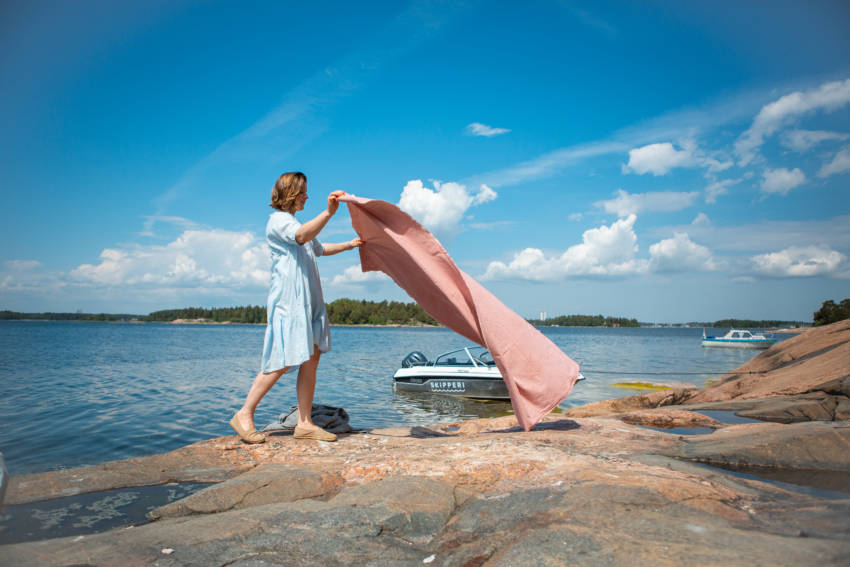 Nainen levittämässä piknikvilttiä rantakalliolle taustallaan vene.